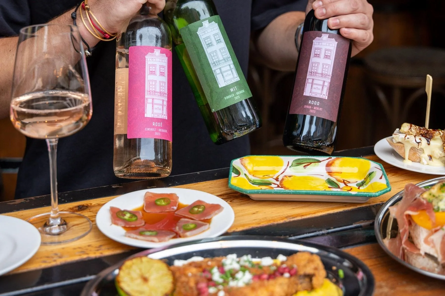 Man houdt drie wijnflessen vast boven tafel met glas rosé en diverse gerechten