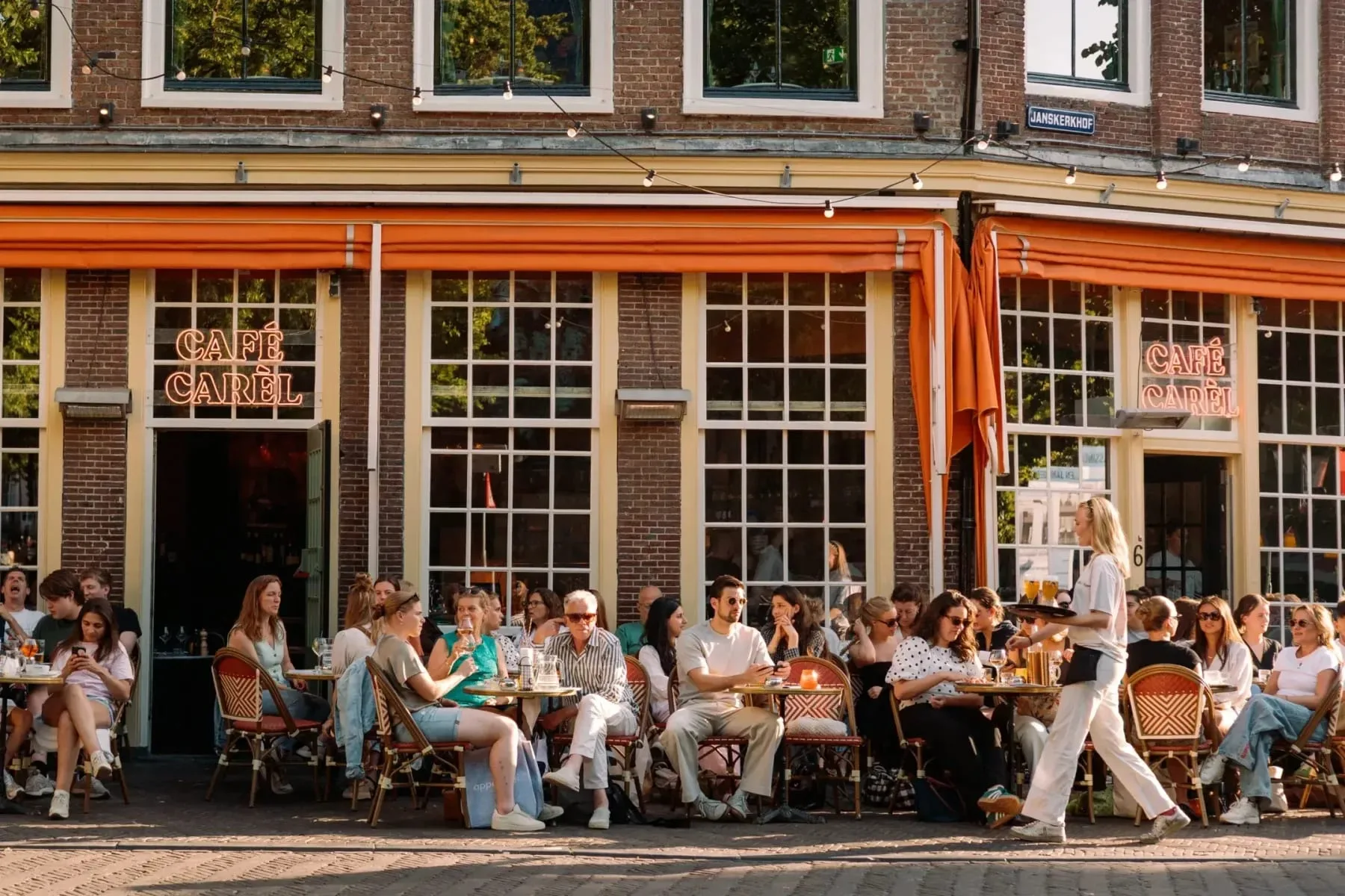 Vol terras met gasten aan tafeltjes voor Cafe Carel onder oranje zonneschermen