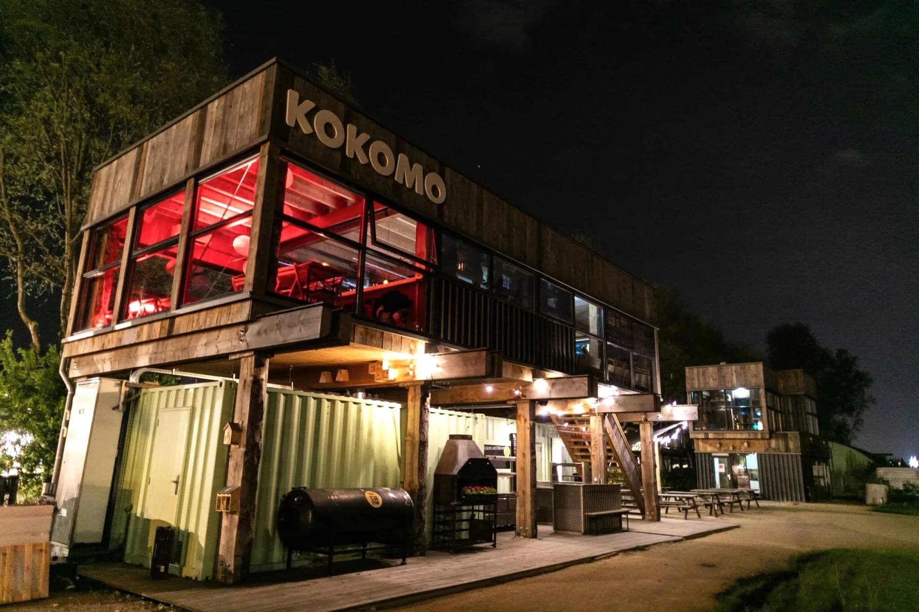 Verbouwde zeecontainers met verlicht Kokomo-logo, rood verlichte bovenverdieping, buitentrap en picknicktafels bij nacht