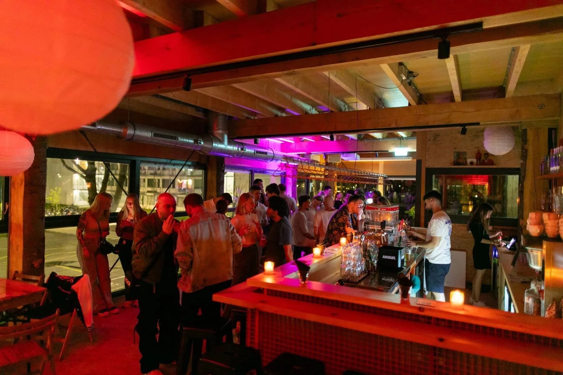 Gasten staan bij de bar in een houten ruimte met roze en rode verlichting