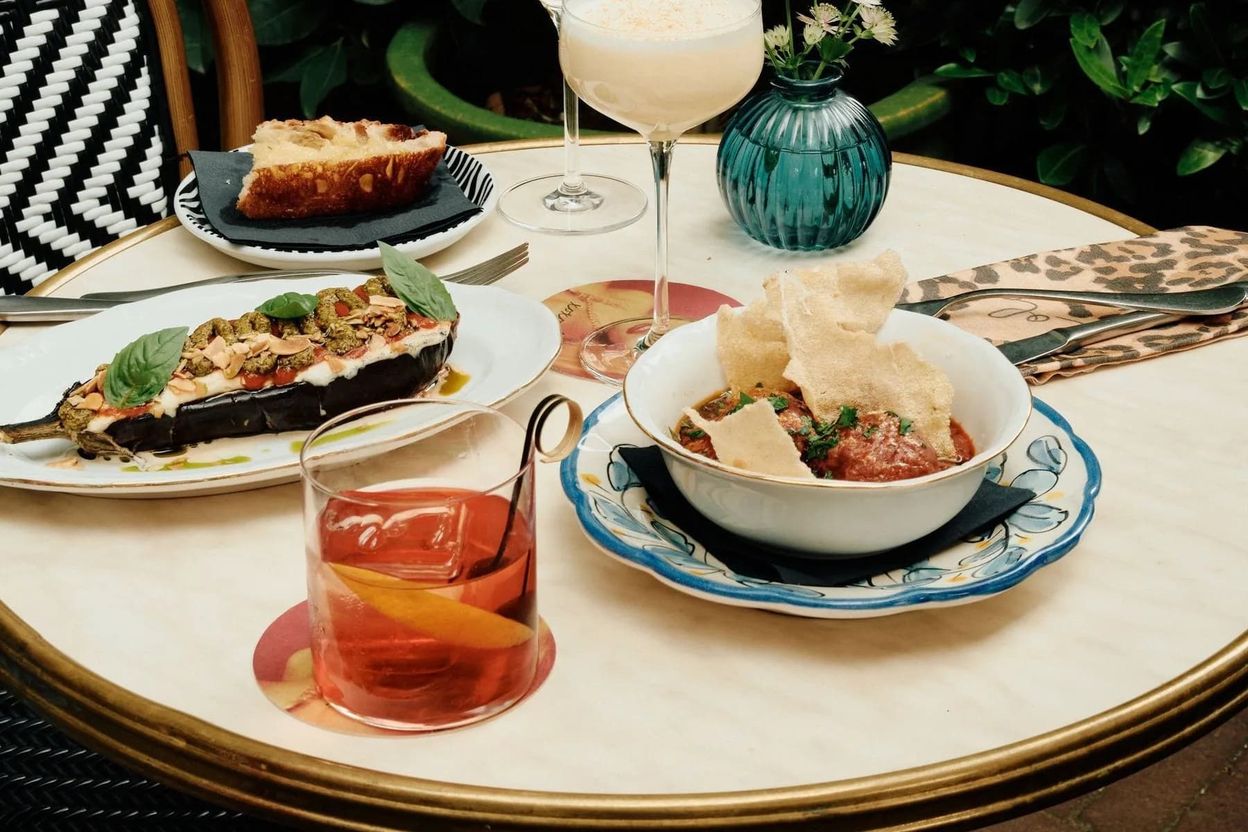 Ronde tafel met gevulde aubergine, brood, cocktails, wijn en bloemenvaasje tussen groene planten
