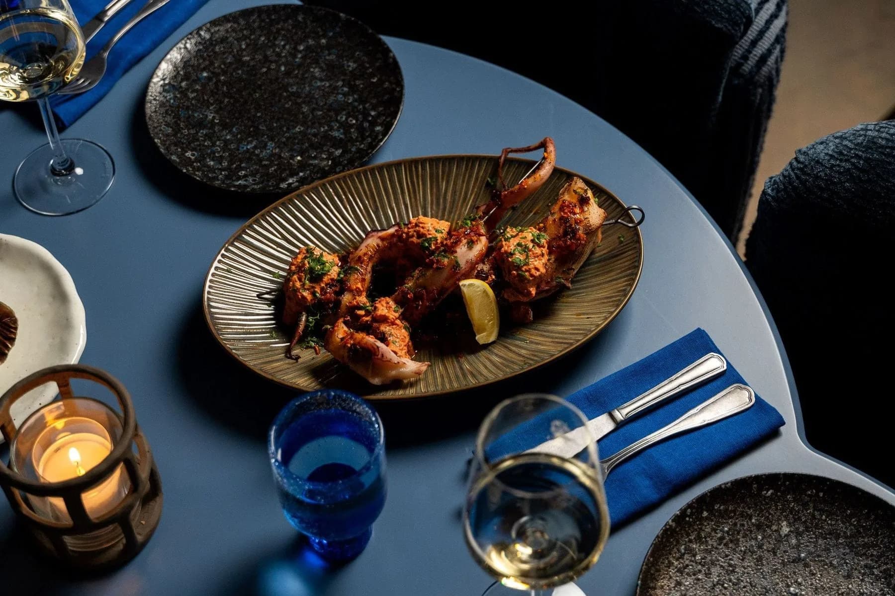 Blauwe tafel met schaal gegrilde inktvis, witte wijn, waxinelicht en donkere borden