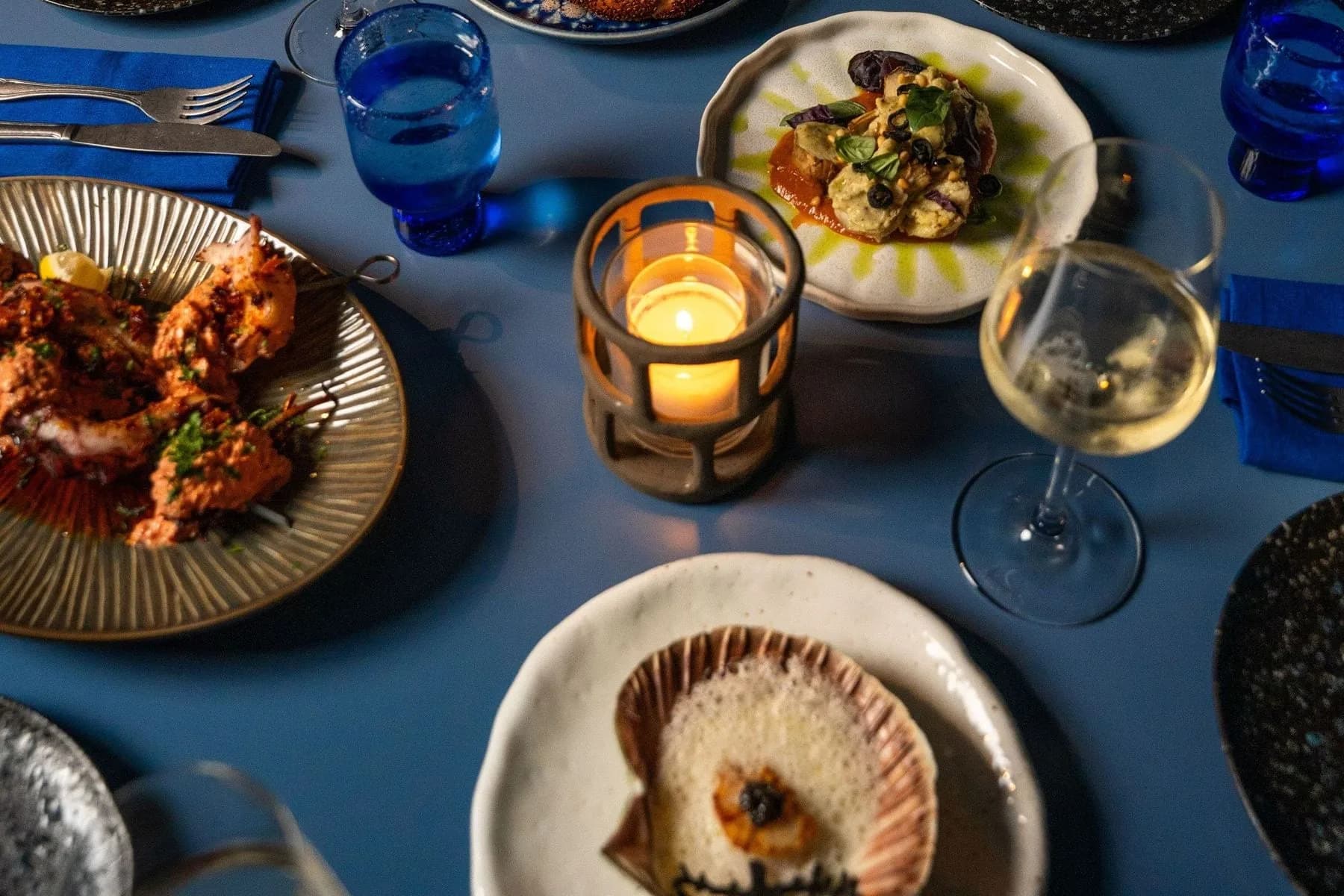 Ronde blauwe tafel met meerdere gerechten, wijnglazen, waxinelichtje en blauwe b