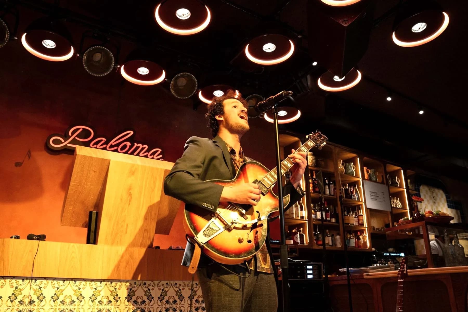 Man met gitaar zingt in microfoon voor bar met flessenwand en neonletters "Paloma"