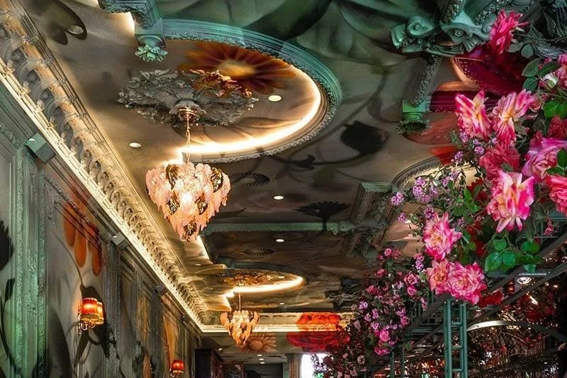 Lange bar met barkrukken, hoge tafels, beschilderd plafond en bloemdecoratie boven de toog