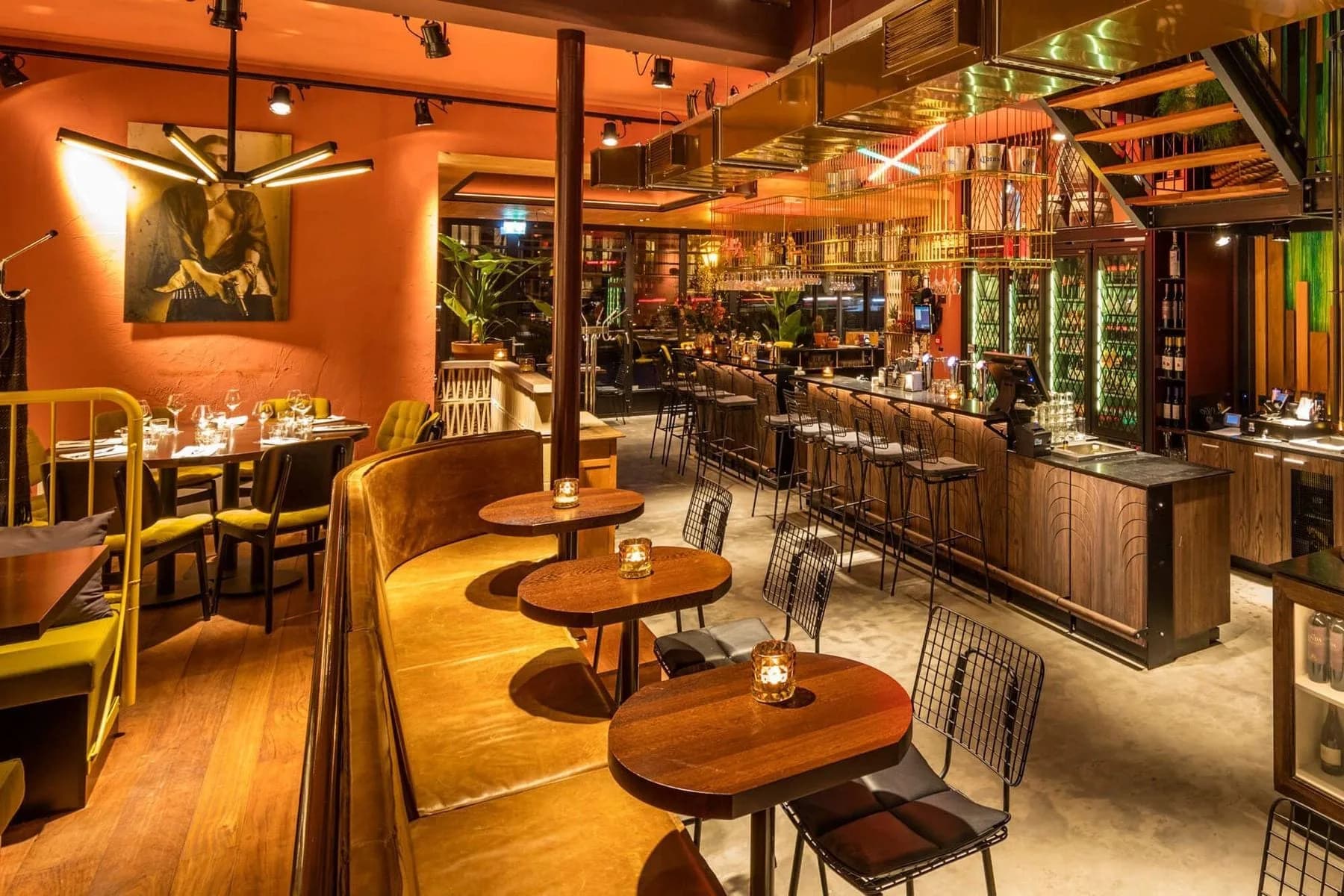 Restaurantzaal met oranje wanden, houten tafels, bankstel en bar met barkrukken