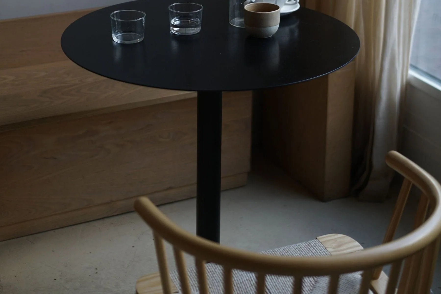 Ronde zwarte tafel met twee koffiekopjes en waterglazen naast houten bank en gordijn