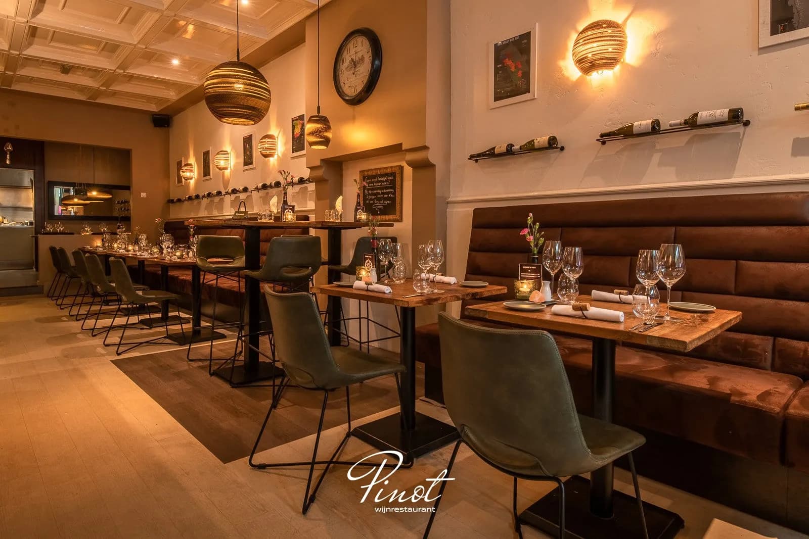Restaurantzaal met houten tafels, groene stoelen, bruine bankstelling langs de muur en hanglampen