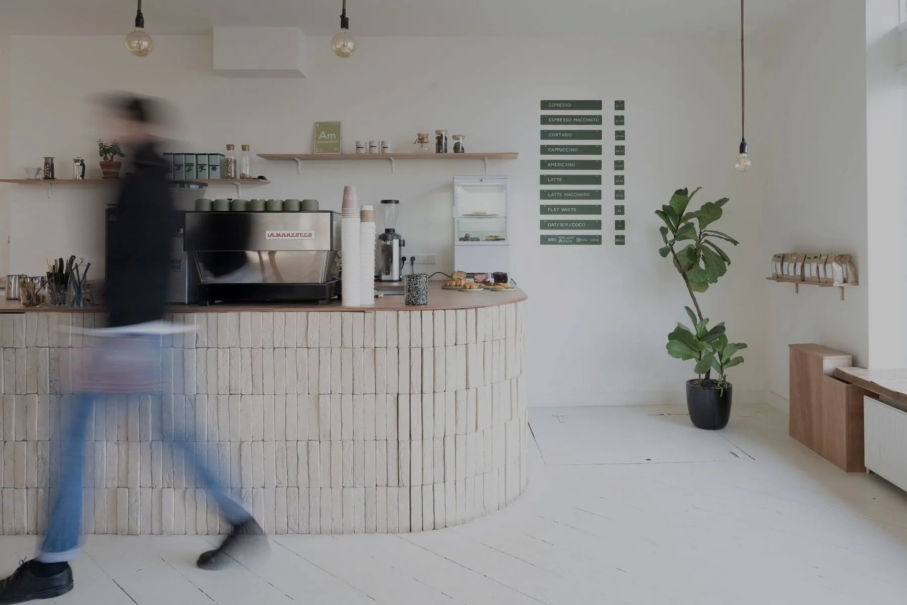 Persoon loopt langs ronde bakstenen toonbank met espressomachine, menubord en plant tegen witte muur