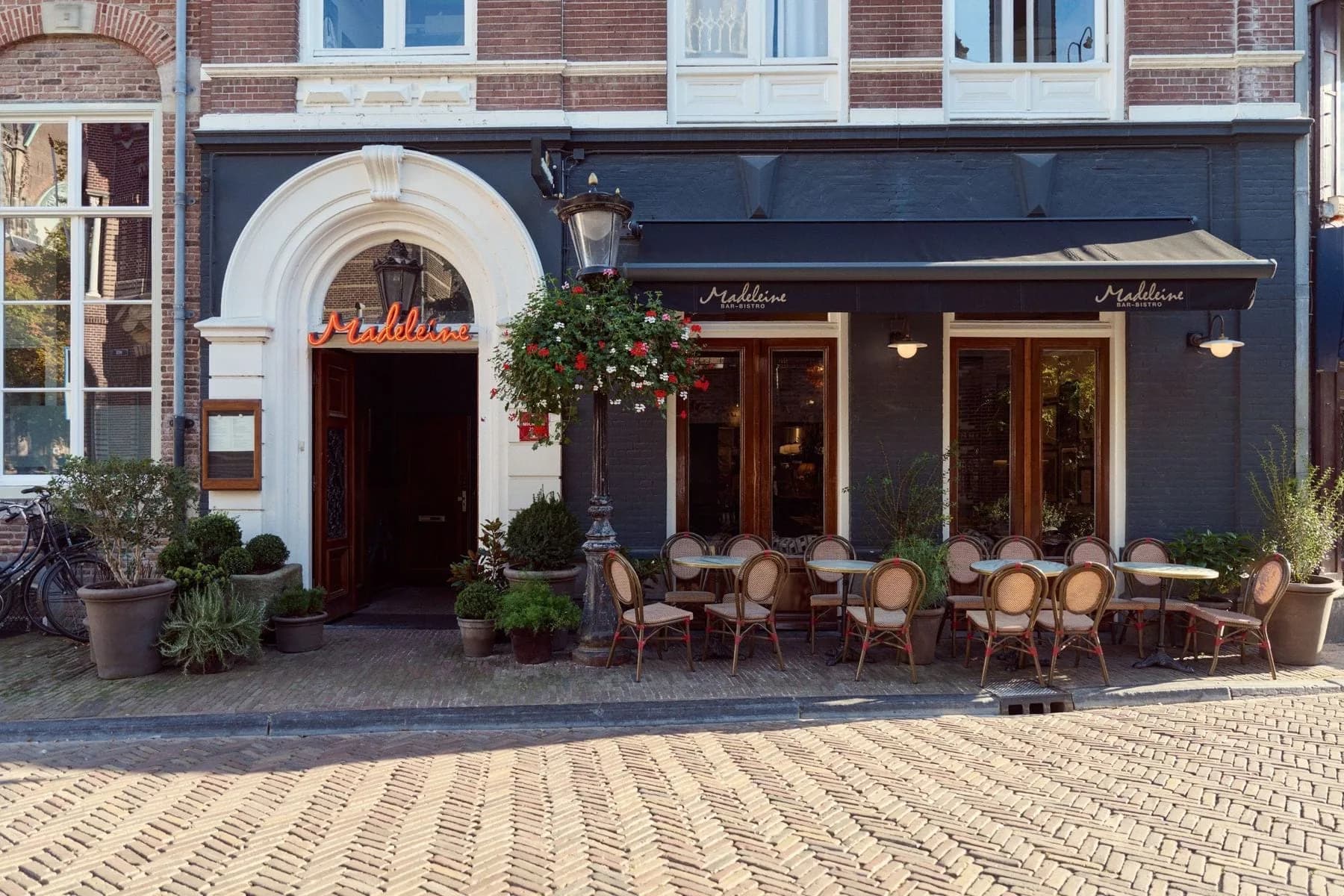 Gevel van Bistro Madeleine met terrasstoeltjes, hangende bloembak en zwarte luifel aan straatzijde