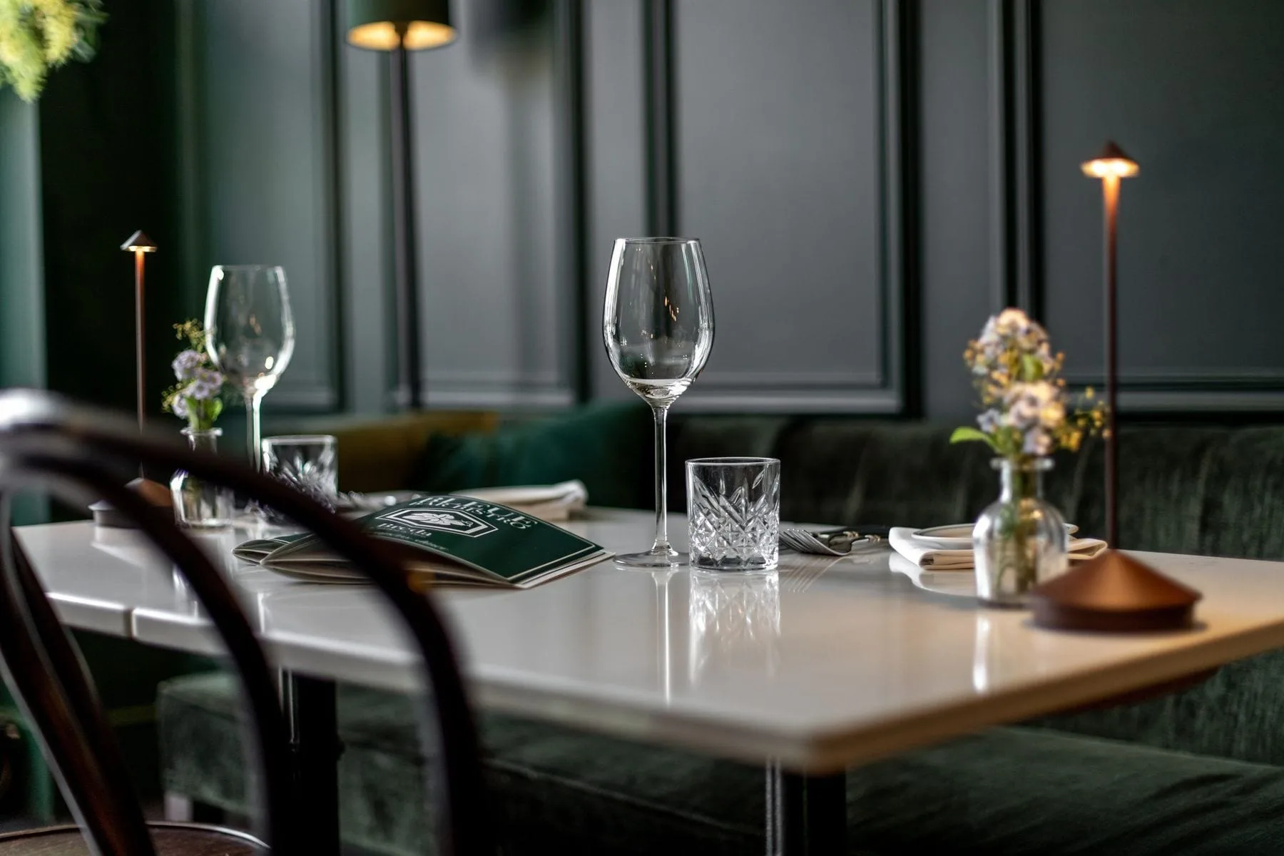 Gedekte marmeren tafel met wijnglazen, menukaarten, tafellampjes en bloemetjes voor groene bank en lambrisering