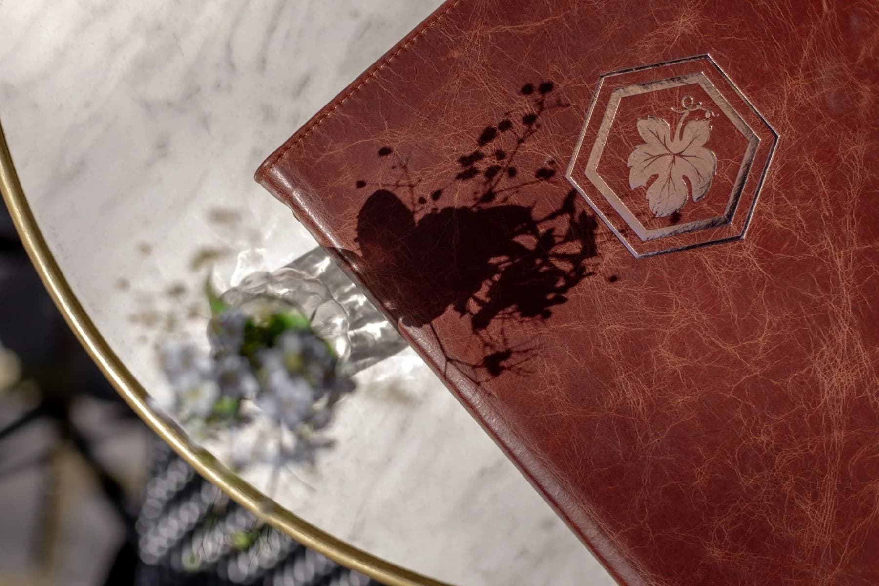 Bruinleren menumap met wijnvlekken en bladlogo op marmeren tafel naast kleine bloemvaas