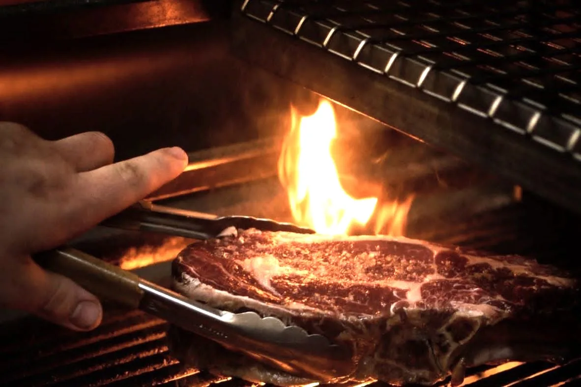 Hand houdt met tang een T-bonesteak boven vlammen op een houtskoolgrill