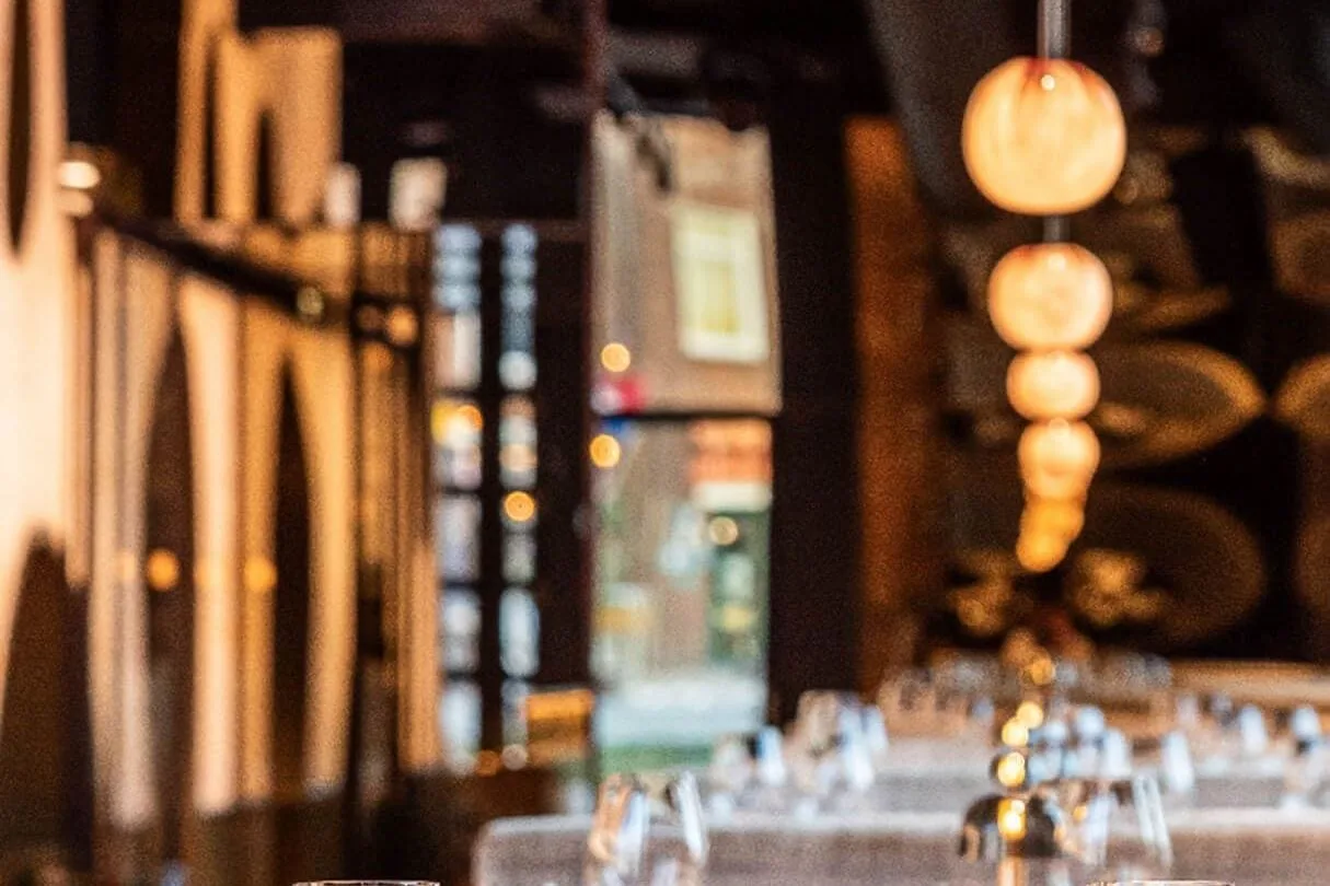 Gedekte tafels met wijnglazen en een klein tafellampje, hanglampen op de achtergrond