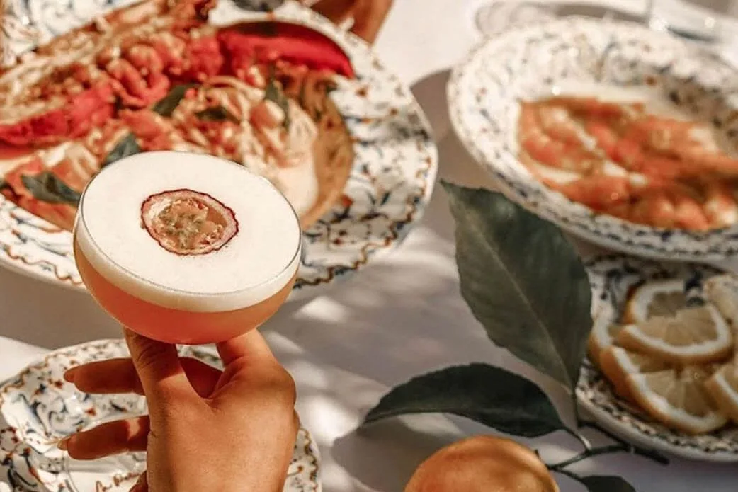 Hand houdt cocktail met passievruchtschijf boven gedekte tafel met kreeftpasta en citroenen