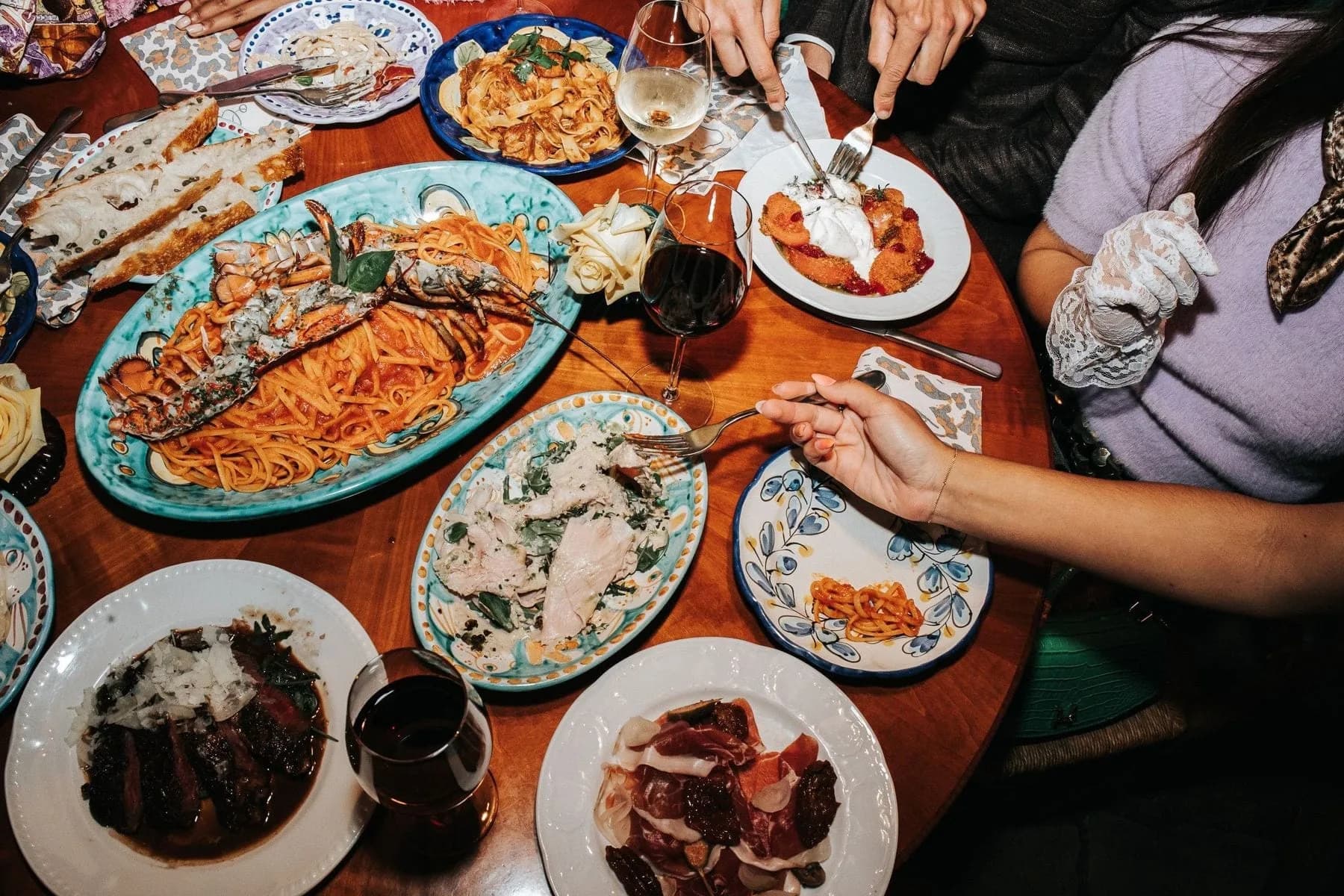 Gasten eten aan houten tafel met borden pasta, kreeft, vlees en glazen wijn