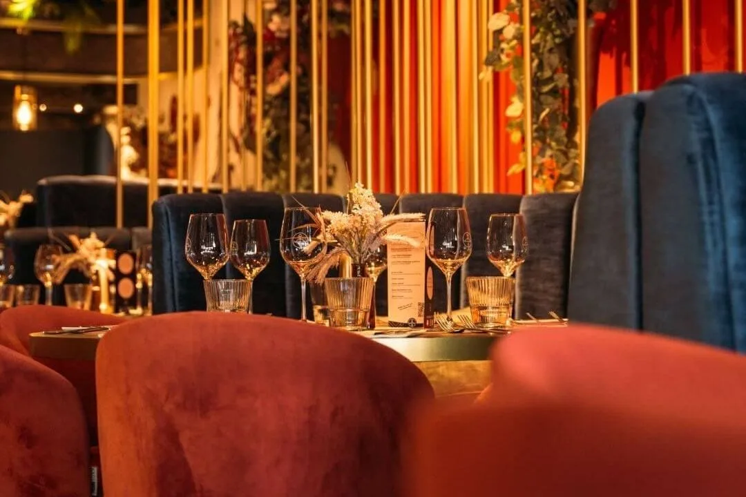 Gedekte tafels met wijnglazen tussen roze en blauwe velours stoelen en gouden scheidingswanden