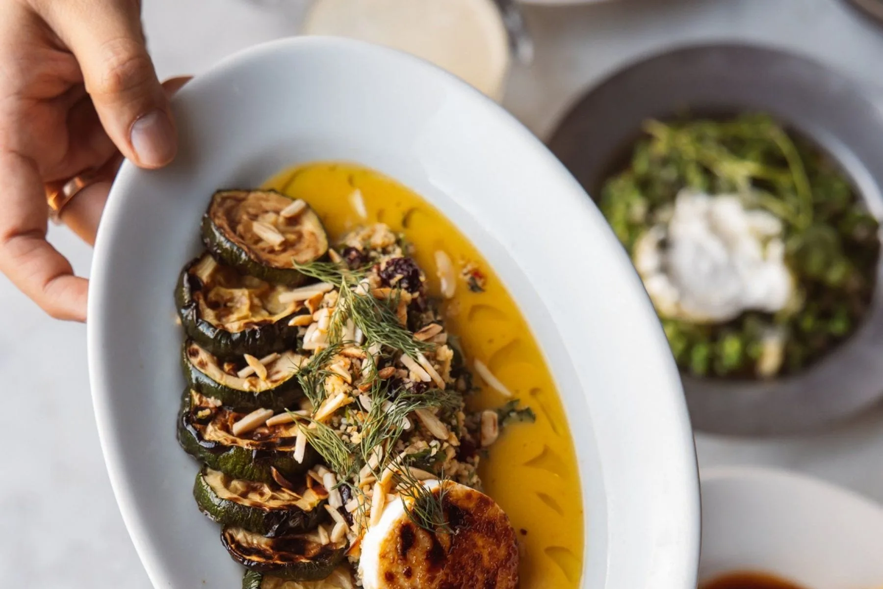 Handen houden een ovaal bord met gegrilde courgette, amandelschaafsel en
