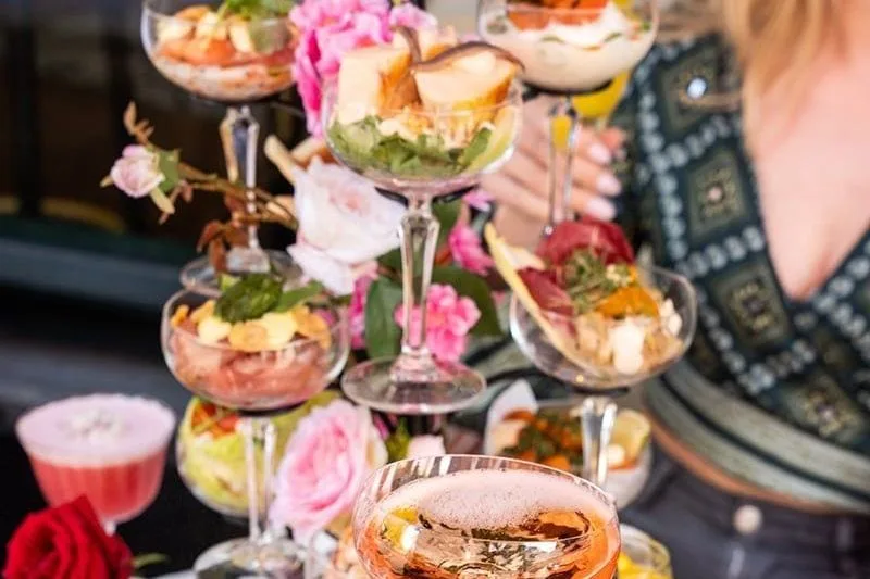 Hand pakt roze cocktail van marmeren tafel met etagère vol hapjes in glazen