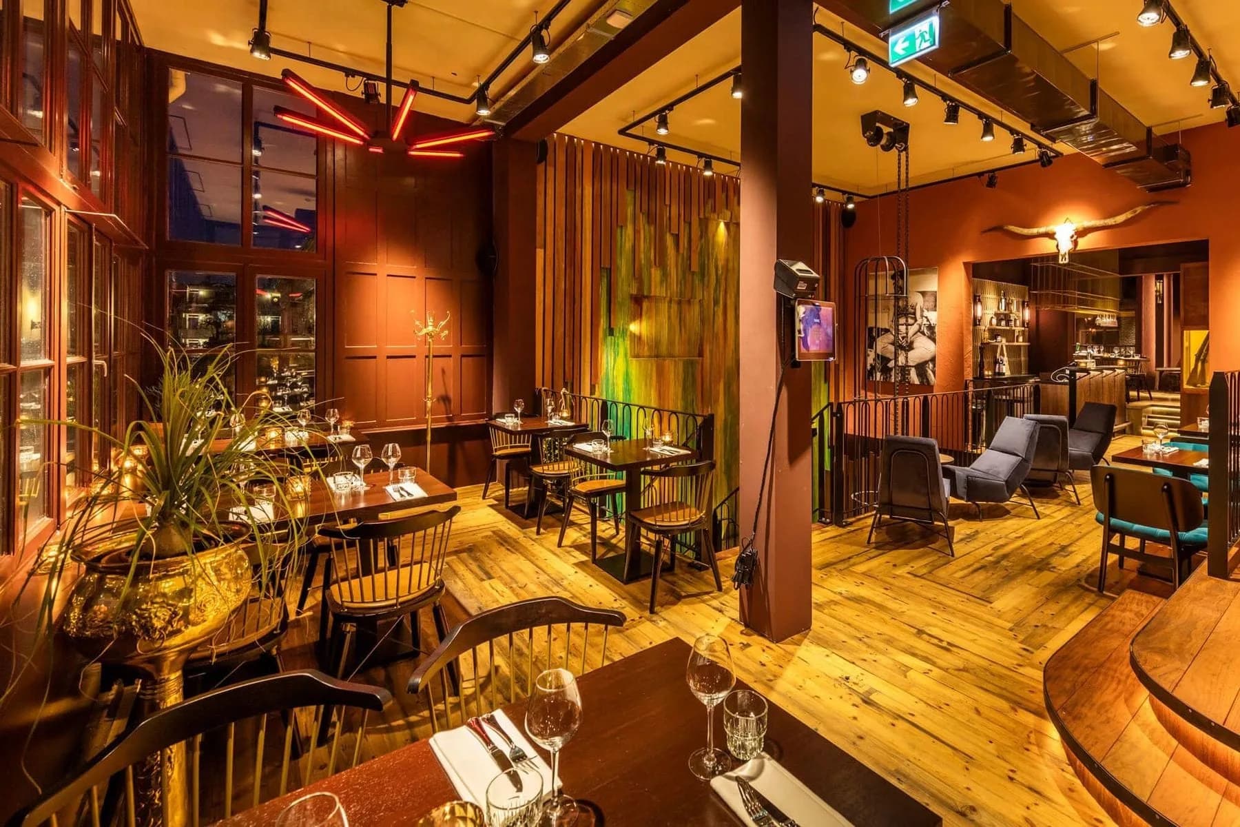 Restaurantzaal met gedekte tafels, houten vloer, spotverlichting aan rails en groene gordijnen