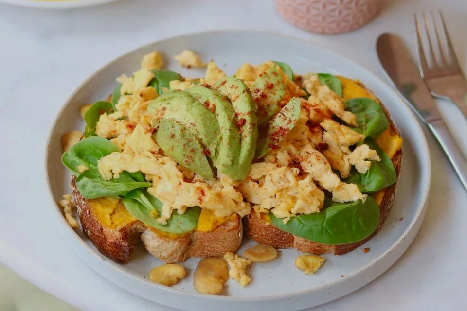 Toast met roerei, avocado, spinazie en paprikapoeder op wit bord naast bestek