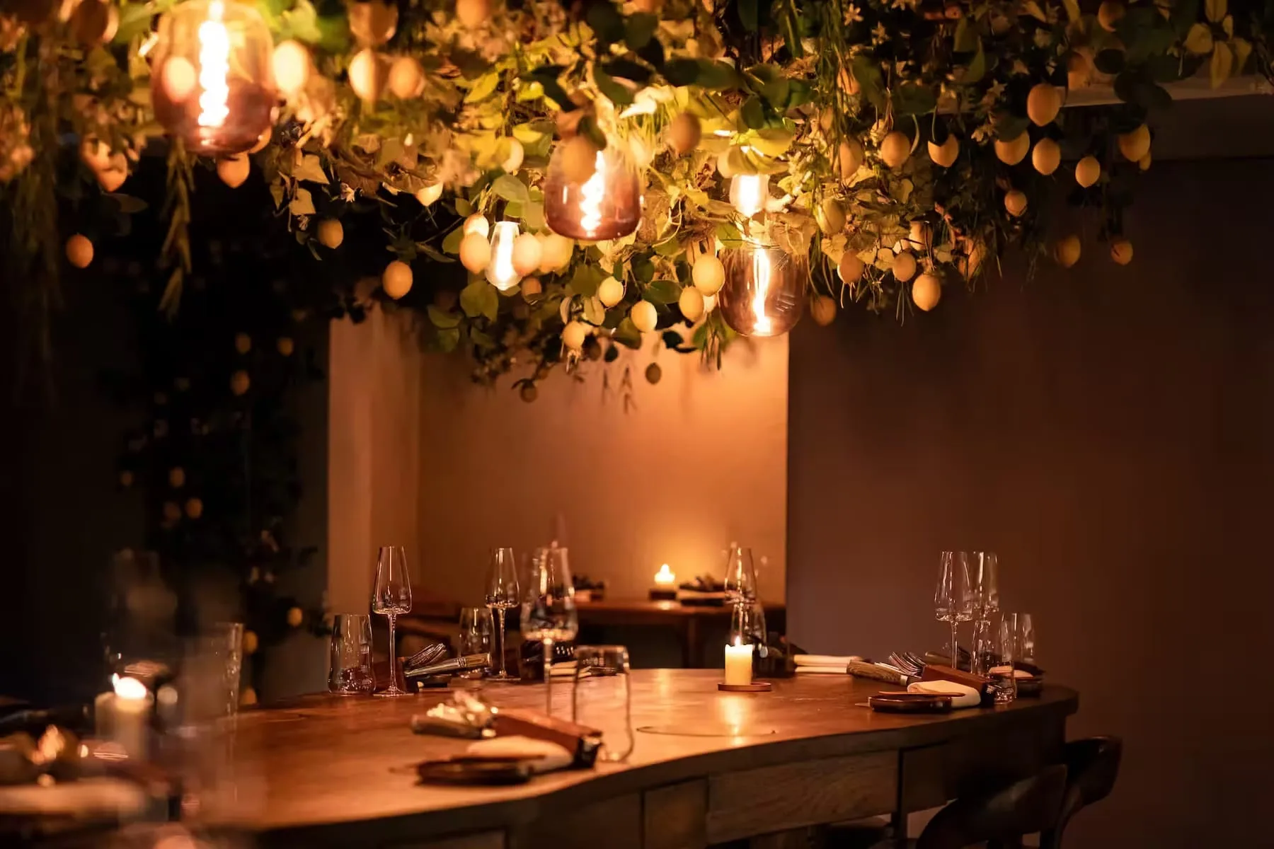 Gedekte houten tafel met barkrukken onder een hangende groendecoratie met citroenen en lampen