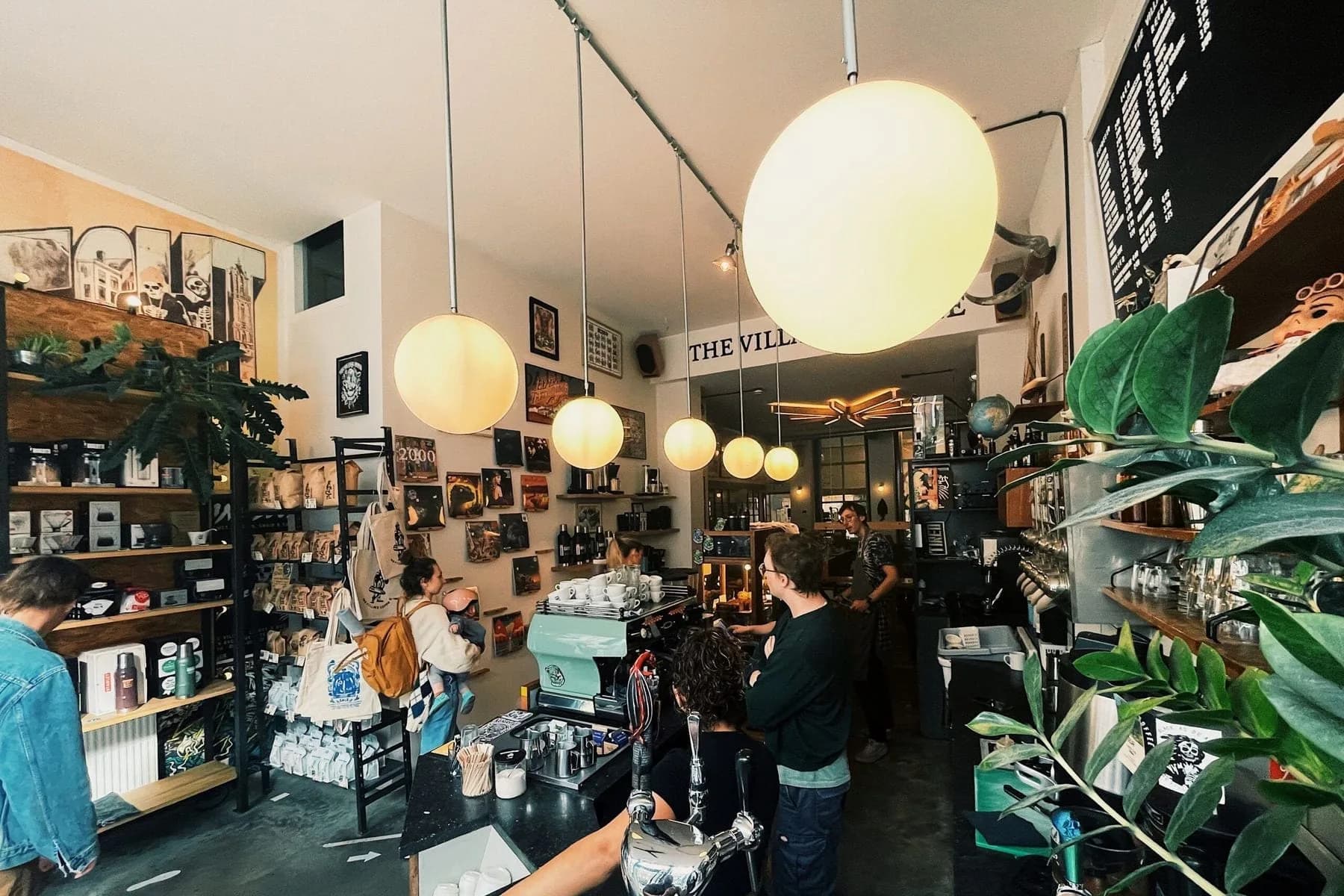 Gasten aan de bar van een koffiezaak met bolvormige hanglampen, planken en planten